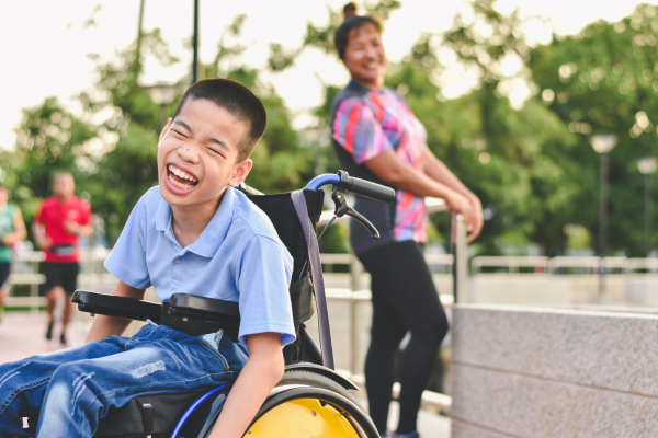 A young child in a wheelchair laughs and smiles in the foreground. A woman in the background who is out of focus looks over at the child with a smile on her face.