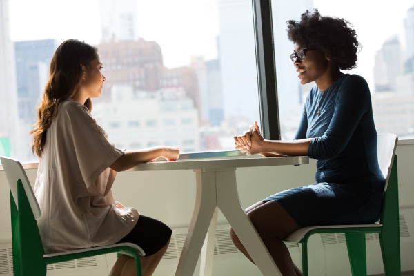 Two women sit at a table by a large window chatting.