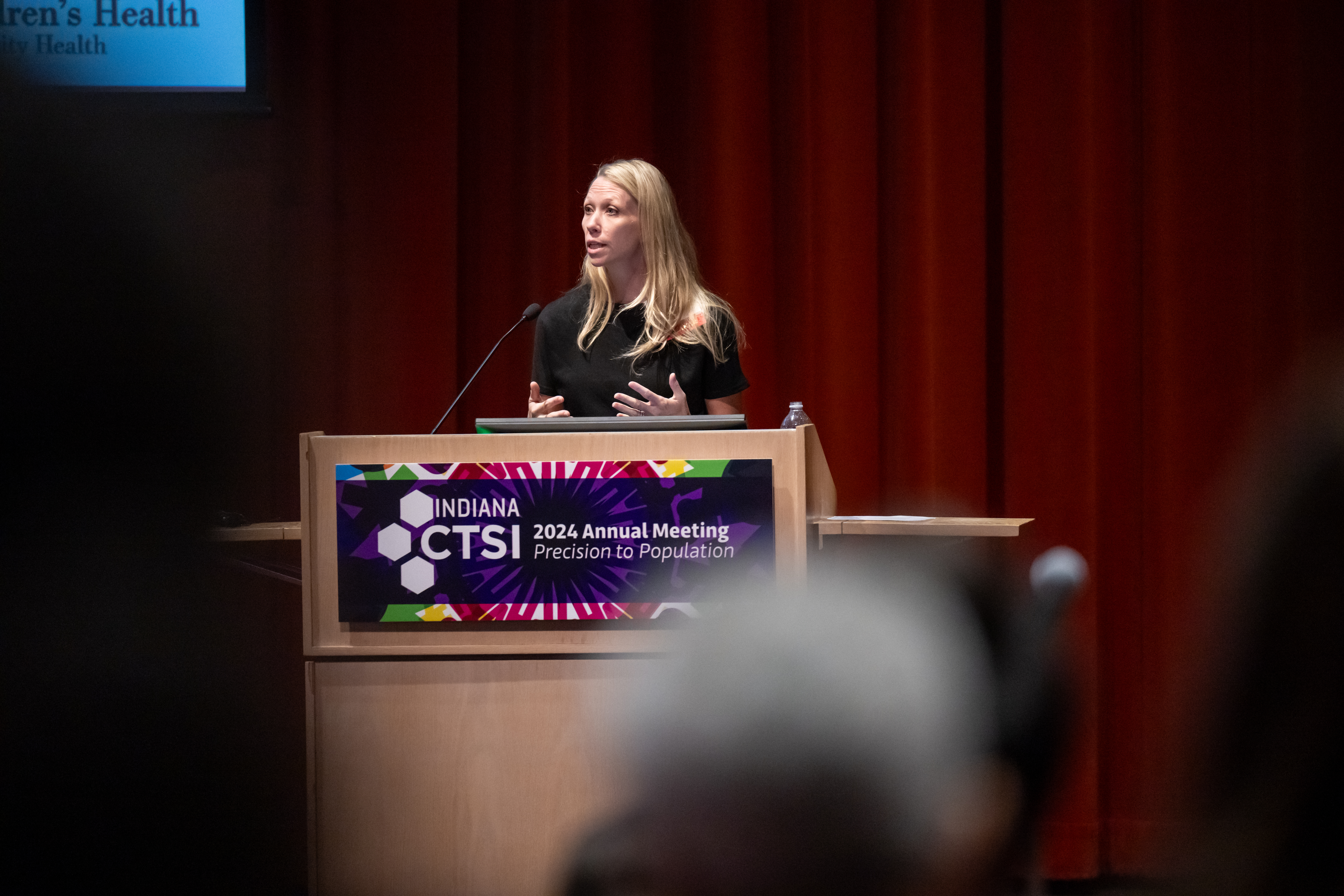 Dr. Rebecca McNally Keehn, a thin white woman with blonde hair wearing a black top, stands behind a podium at the 2024 Indiana CTSI Annual Meeting. She is presenting her work and stands in front of heavy red drapes.