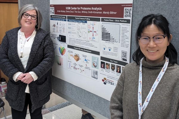 Two IU School of Medicine Proteome Analysis Core members stand smiling in front of a poster detailing their available services.