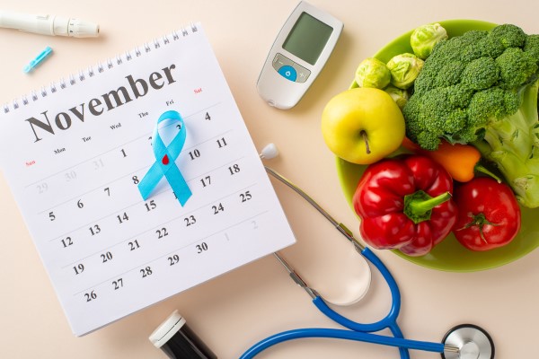 a calendar lays on a table top surrounded by a bowl of brightly colored fresh vegetables, a blue and silver stethoscope, a sugar monitor, and other diabetes related supplies. The calendar page is open to November, and a blue ribbon is pinned over the dates.