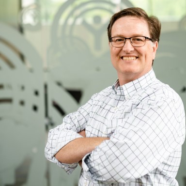 Jonathan Barclay smiles in a headshot. He is a Caucasian presenting man with short brown hair, and glasses. He is wearing a white checkered button up, and stands with his arms folded across his chest in front of a glass backdrop.
