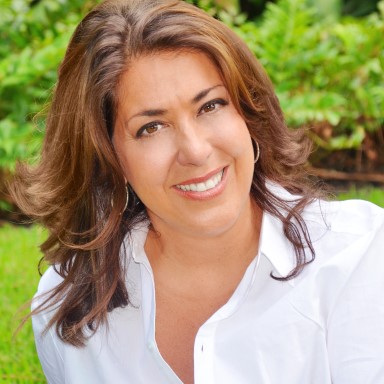 Carla Heiser smiles in a headshot. She is wearing a white button up shirt, and has medium brown hair and brown eyes. She is against a background of lush green vegetation.