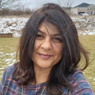 Nasreen Lalani smiles in a headshot. Her long brown hair is windswept, she wears a blue and red plaid shirt on a background of slightly snow covered grass and landscape.