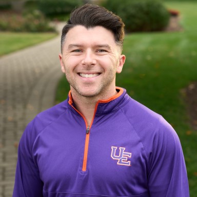 William Miller smiles in his headshot. He is a young Caucasian presenting man with short brown hair and a wide smile. He wears a purple and orange partial zip shirt with the letters UE on the left pectoral.