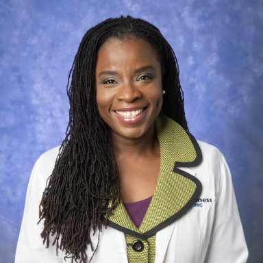 Pearl Quartey smiles in her headshot. She is an African American woman with black hair, dark eyes, and a wide smile. She wears a white doctor's coat over a green blazer and purple top in front of a blue background.