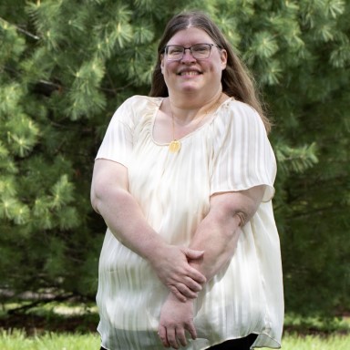 Jennifer Stefancik poses for a headshot. She is a Caucasian presenting woman with long dark blonde hair, glasses, and a smile. She wears a white flowy blouse with her arms crossed across her body in front of a background of lush green vegetation.