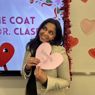 Suparna Clasen smiles as she holds a pink paper heart that says behind the coat.