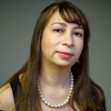 Lupita Valdes poses in a headshot. She has long medium brown hair with bangs that sweep across her forehead, and wears a strand of pearls around her neck with a black sleeveless top against a grey background.