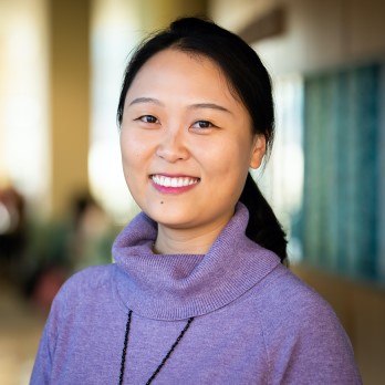 Haocen Wang poses in a headshot. She is an Asian woman with black hair that is secured in a ponytail and dark eyes with a wide smile. She wears a purple turtleneck style top.