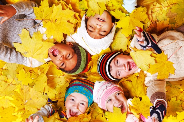 a group of diverse children in various hats lay in a pile of bright yellow leaves, making various expressions of joy