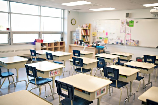A classroom is set up in a room with large sunny windows in the upper left corner. There are student desks organized in rows facing the teacher's desk and a large white board to the upper right hand corner of the image.