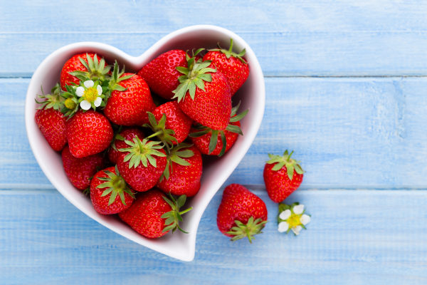 A white heart shaped bowl filled with bright red strawberries sits on a blue wood grain backdrop. There are two strawberries and a strawberry flower laying on the table next to the bowl.