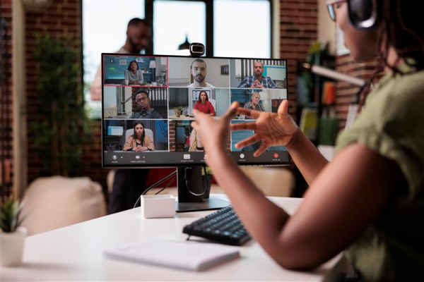 A woman appears to be speaking on a web conference. There is a computer monitor in the background of the image with multiple people on camera.