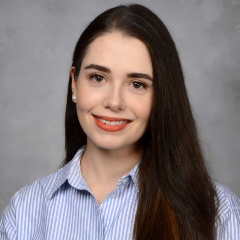 Daniela smiles in her headshot. She is a younger woman with fair skin and dark hair. She wears a blue and white striped button up