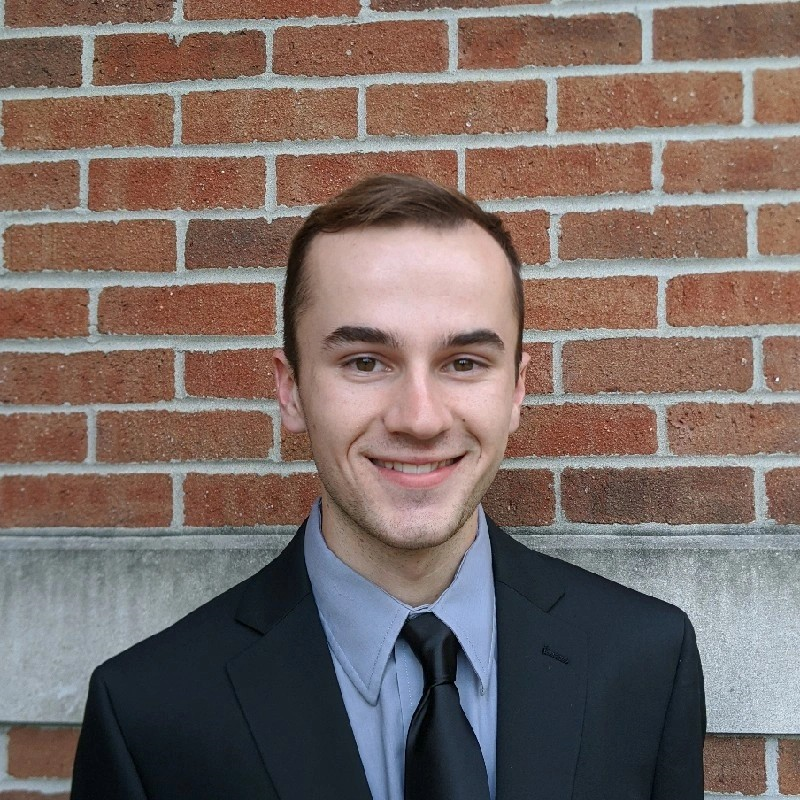 Luke smiles in his headshot. He is a younger white man with closely cropped brown hair. He wears a cornflower blue button up shirt under a black suit jacket with a black tie, and stands in front of a red brick wall.