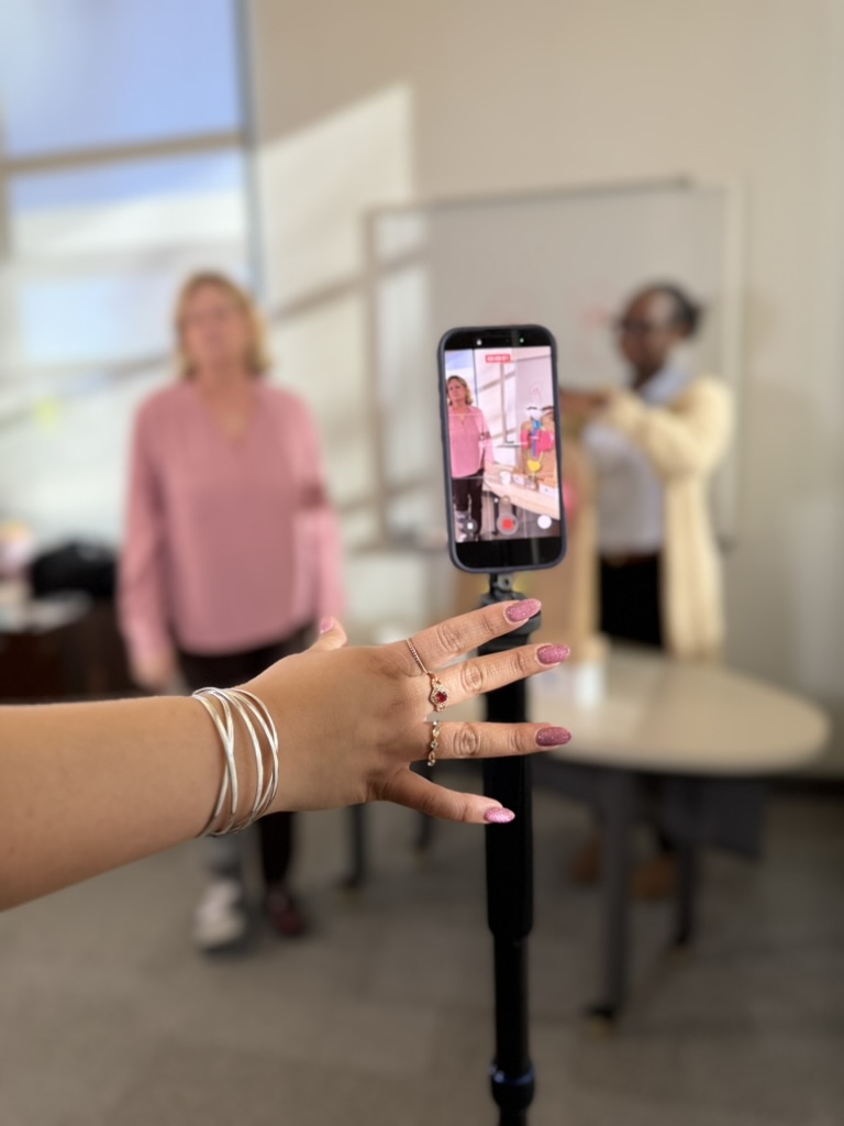 The scene with Sharon Moe and Fatma Niang as seen through an iPhone camera. Bonnie's hand on the phone stand, with jewelry and pink manicure, visible.