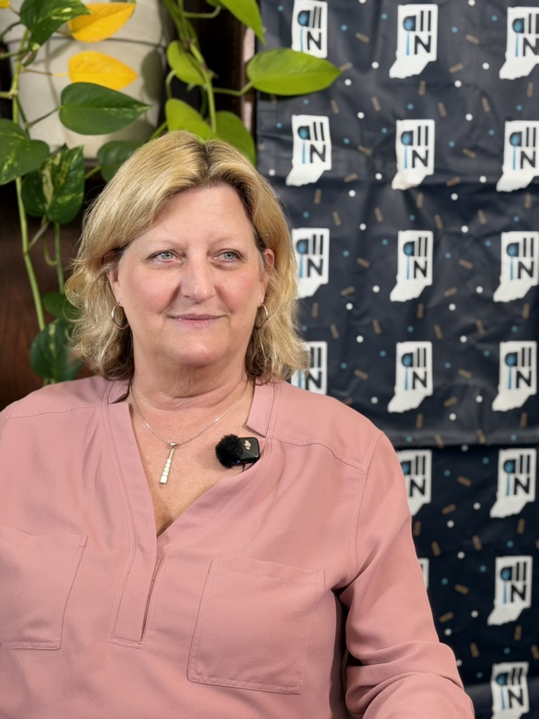 Portrait of Sharon Moe, seated, against a backdrop of All IN logos and a vining potted plant. 