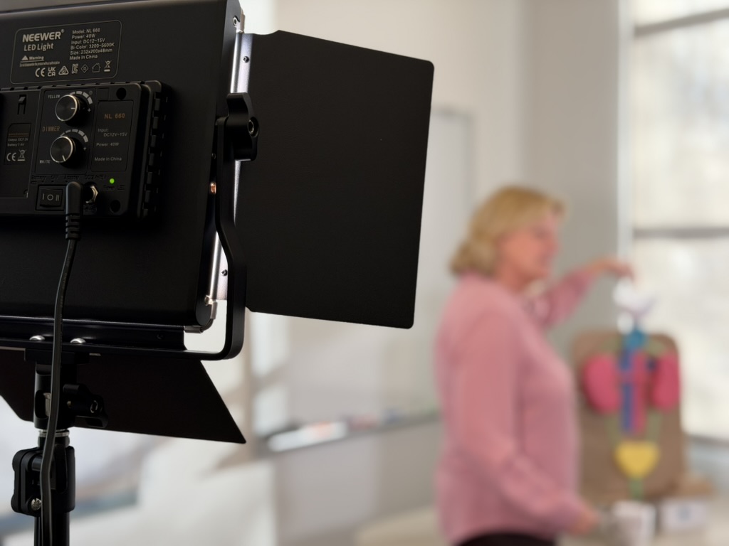 Closeup of the back of a light, in focus. Out of focus, Sharon Moe while being filmed.
