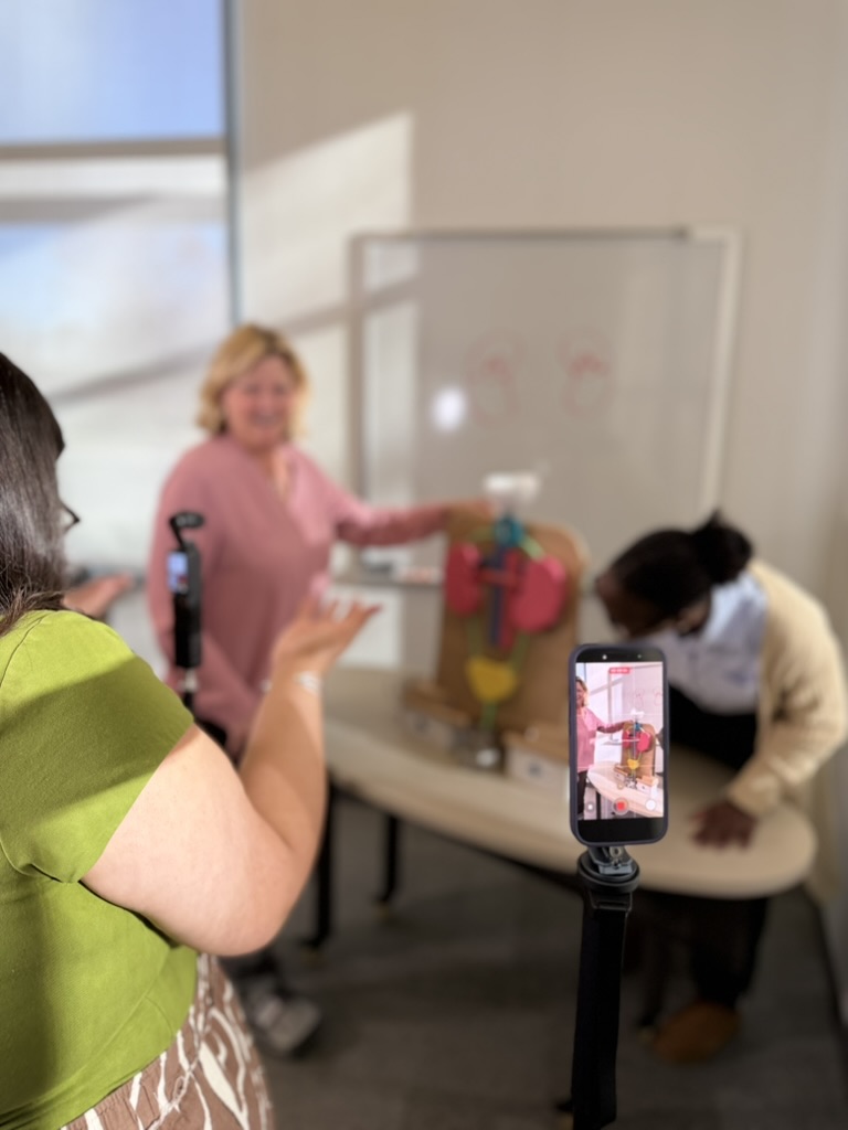 Sharon Moe and Fatma Niang are filmed working with the kidney demo model. Bonnie is behind one camera and the scene is visible through the phone camera. 