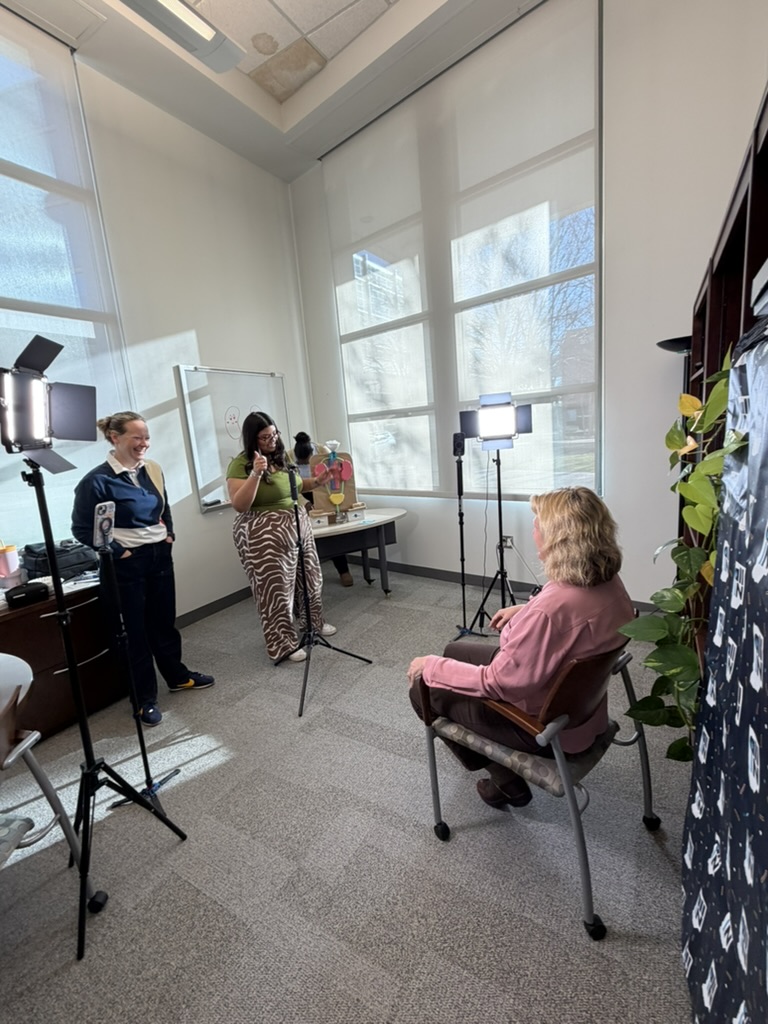 Video shoot set-up in a brightly lit office. Sharon Moe seated speaking with Lynsey Delp and Bonnie Marcelo.