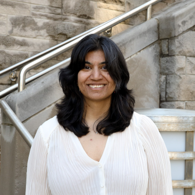 Natasha Chaku, PhD, smiles in her headshot. She wears a white button up blouse and stands in front of a stone wall and stairs.