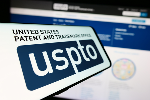 The US Patent website is unfocused in the background while a phone is held in the foreground with the words "United States Patent and Trademarks Office uspto" on it.