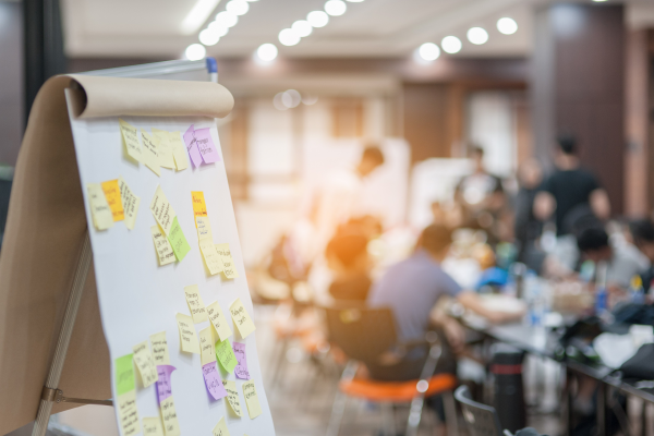 A workshop with numerous attendees appears to be taking place in the background, with a white paper chart covered in colorful post it notes in the foreground.