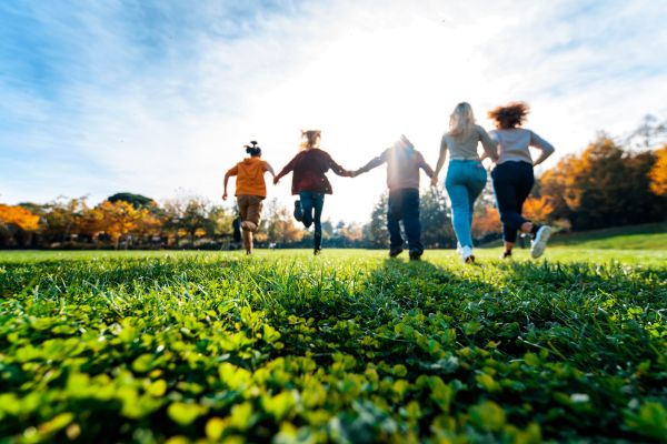 A line of children frolic in a green field with fall foliage in the background.
