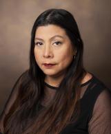 Headshot of Adriana Hung, MD, MPH, of Vanderbilt University Medical Center. Hung is seen wearing a black top that is opaque on the shoulder straps and chest, and mesh material on the upper arms and collar. Hung's hair is long and dark, and they are looking into the camera with a soft, closed-mouth smile. The background behind Hung is a grey/brown backdrop.