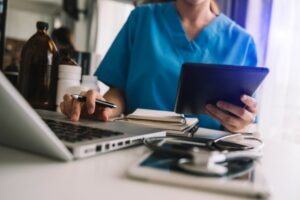 Image of a person in scrubs working on a laptop while also holding a tablet, surrounded by medical tools