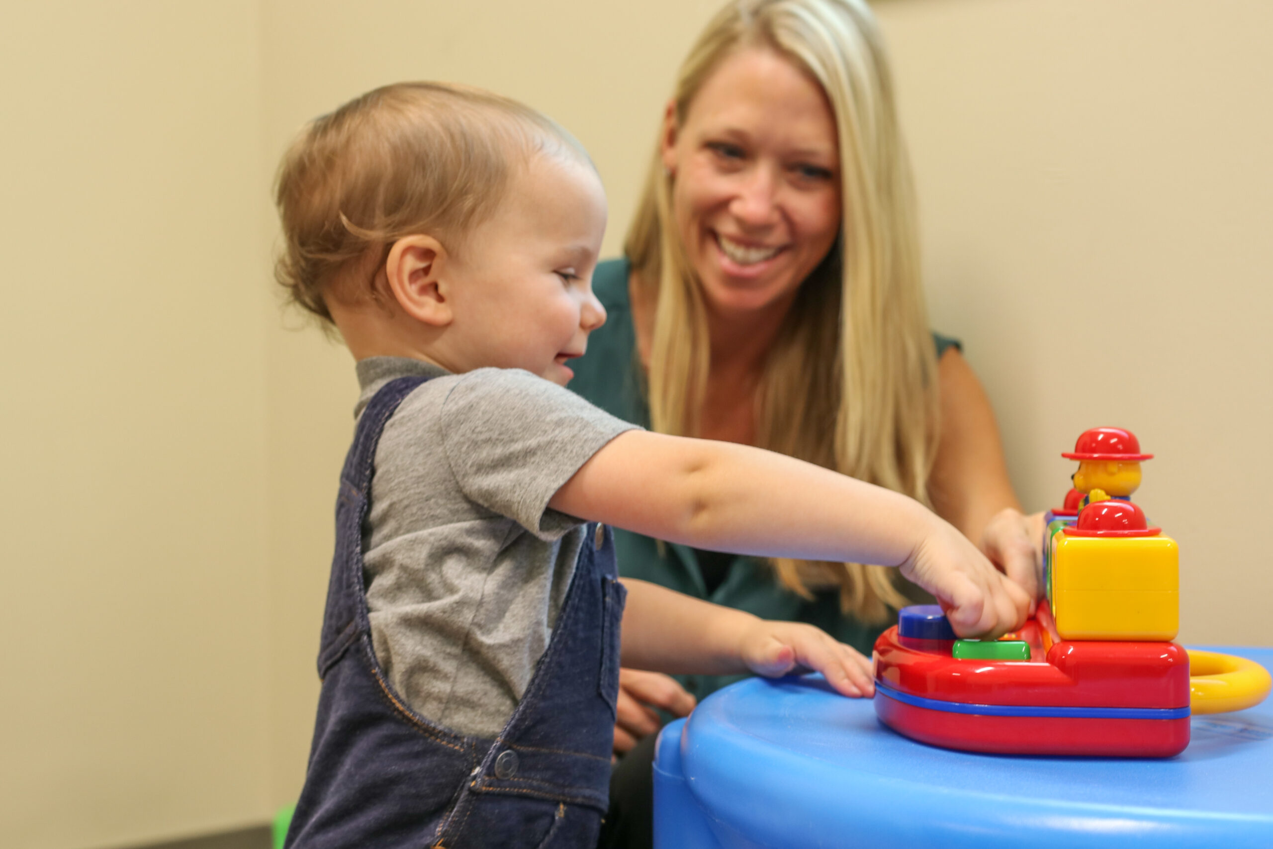 Dr. Rebecca McNally Keehn, a slender white woman with blonde hair, smiles in the background as she watches a toddler in the foreground with a grey shirt and denim overalls interact with a colorful toy on a blue table.