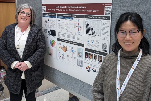 Two IU School of Medicine Proteome Analysis Core members stand smiling in front of a poster detailing their available services